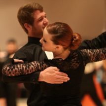 Two smiling ballroom dancers in black perform their routine.