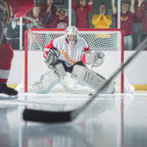 A hockey goalie attempts to block a shot as fans cheer on behind the safety glass in the background.