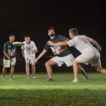 Four men playing ultimate frisbee, a player in a red hat barely gets a throw past a defender wearing white.